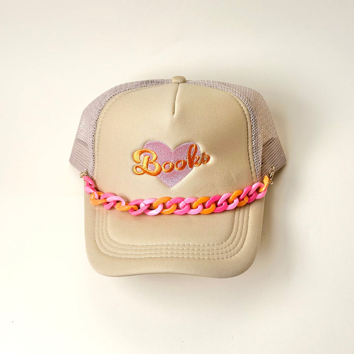 The Darling Effect's beige mesh trucker hat features a heart and "Books" embroidered on the front, accented with an Acrylic Sassy Trucker Hat Chain in pink and orange across the brim. The hat is shown on a plain white background.