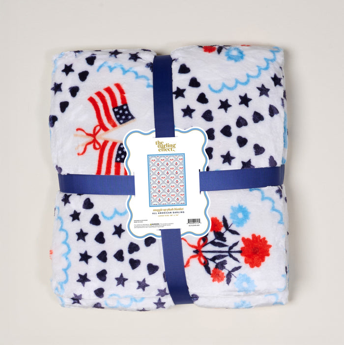The Darling Effect’s All-American Darling Snuggle Up Plush Blanket features white plush fabric with blue, red, and navy stars, hearts, flowers, and American flags—finished with a blue ribbon. Cozy up anytime with this super-soft blanket.