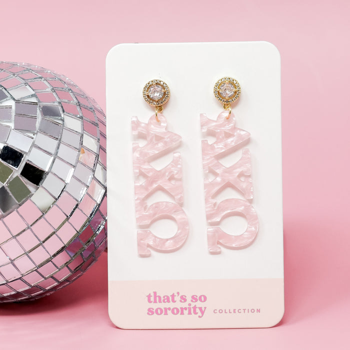Alpha Chi Omega Letter Pink Acrylic Earrings by The Darling Effect are shown on a white "that's so sorority collection" card, beside a silver disco ball on a pink background—perfect for sisterhood events or bid-day baskets.