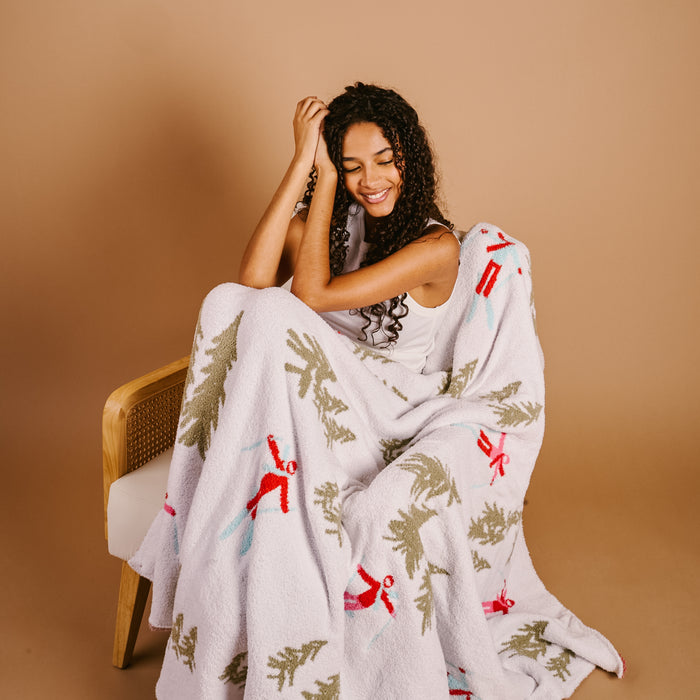 A woman with curly hair sits on a chair, smiling with her eyes closed, wrapped in The Darling Effect's Aprés Ski Cozy Dreams Luxe Blanket featuring green trees and red stick figures on a beige background.