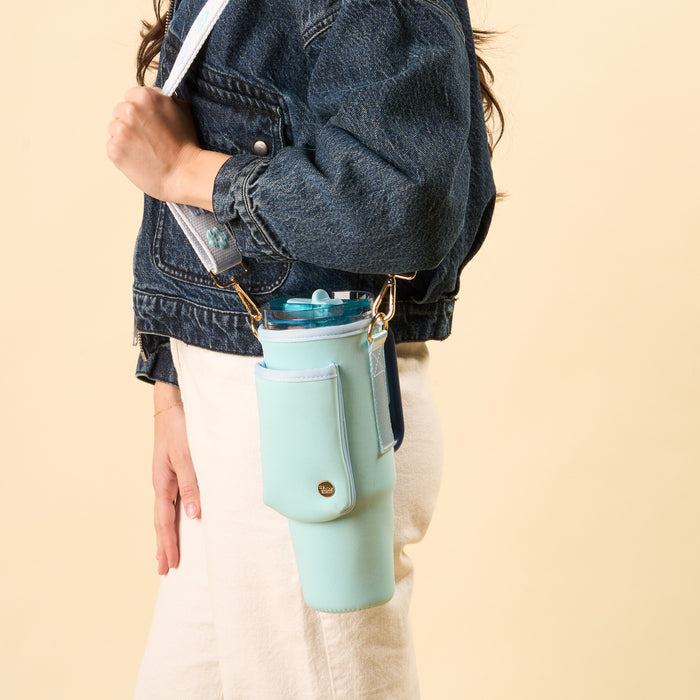 A person in a denim jacket and light pants carries the-darling-effect’s Aqua Tumbler Sling with an adjustable strap, holding a pastel blue tumbler hands-free against a beige background.