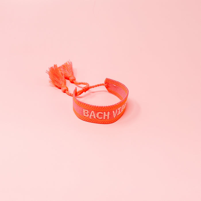 The Darling Effect's Bach Vibes Woven Bracelet features pink adjustable weaving, playful tassels, and "BACH VIBE" in white embroidery. Shown on a pink background, it's perfect for stacking with your favorite trendy bracelets.