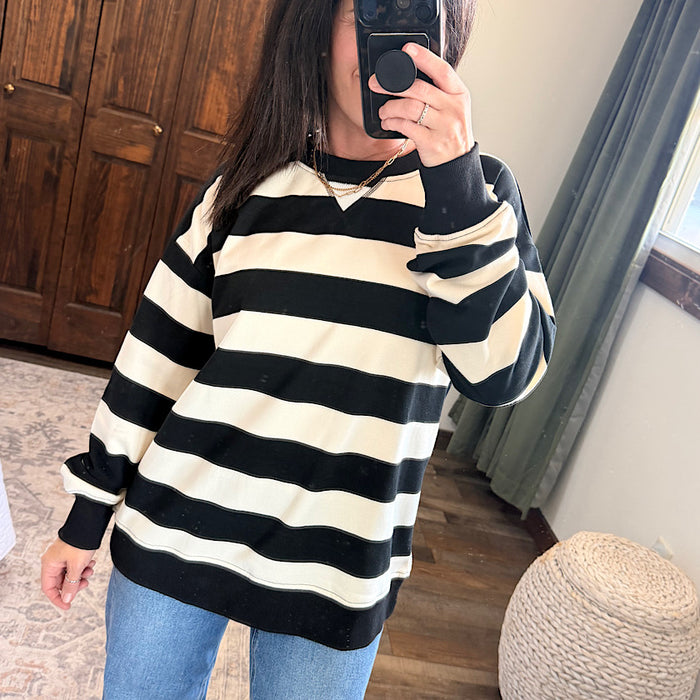 A person taking a mirror selfie wearing a black and cream horizontal striped long-sleeve sweatshirt with a crew neck, paired with blue jeans in a home setting.