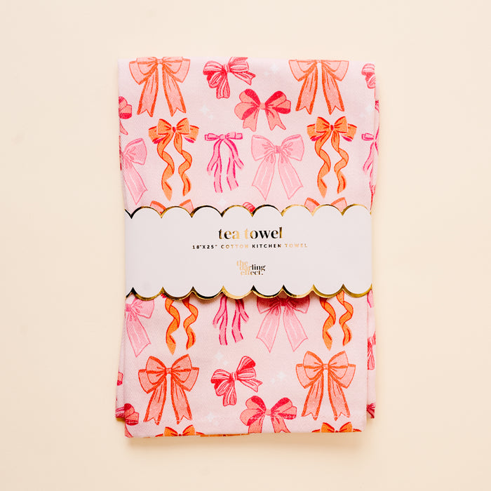 The Blushing Bows Tea Towel by the-darling-effect features a pink and orange bow pattern, folded and wrapped in a white paper band labeled “tea towel” and “18x25 cotton kitchen towel,” shown on a light beige background.