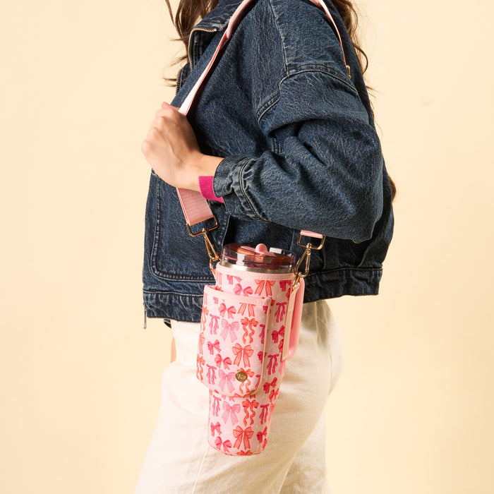 Against a beige background, someone in a denim jacket and white pants carries the-darling-effect's Blushing Bows Tumbler Sling—a pink, patterned, hands-free tumbler holder with an adjustable strap. The person's head is not shown.