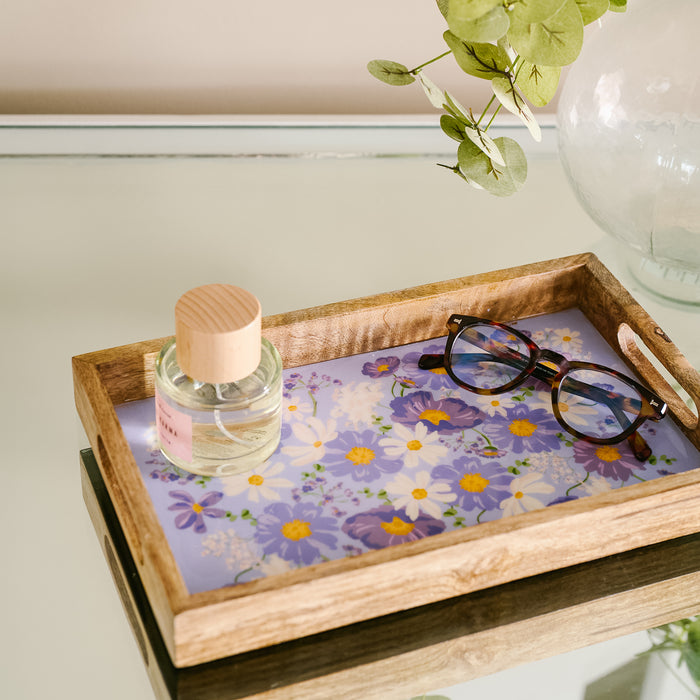 The Bouquet Beauty Purple Rectangle Wood Enamel Tray by the-darling-effect rests on a reflective surface, displaying a perfume bottle and tortoiseshell glasses amid purple and white floral accents.