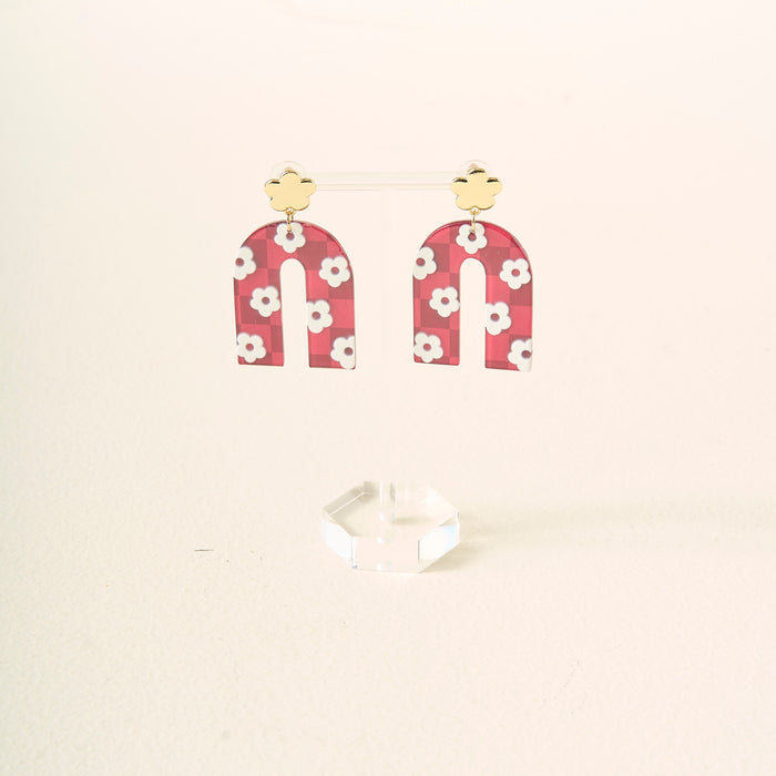 A pair of burgundy, arch-shaped acrylic earrings with white flowers and gold stud tops by The Darling Effect, displayed on a clear acrylic stand—perfect for showing off your school colors.