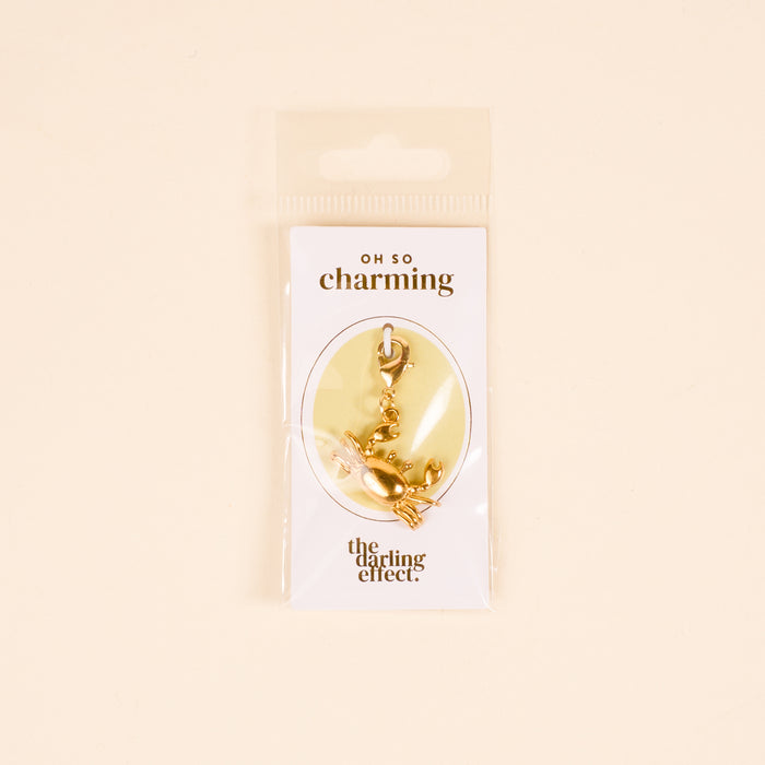 The Darling Effect’s Crab Jewelry Charm Gold hangs from a clasp on a white “OH SO charming” card, perfect for your charm bar or jewelry collection. The card comes in clear plastic packaging.