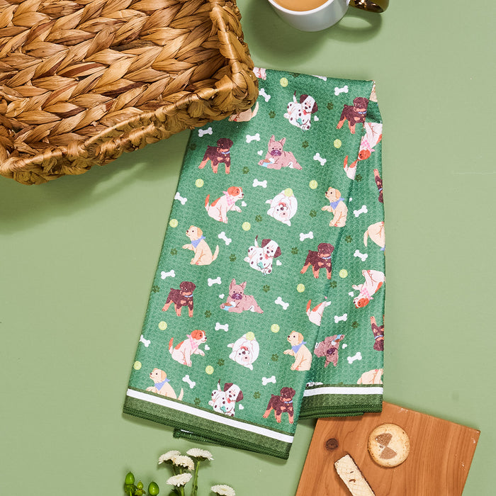 The Darling Effect’s Dog Days Waffle Tea Towel, adorned with playful cartoon dogs and white bone patterns, is displayed on a green surface beside a wicker basket, tea cup, and wooden board with cookies.