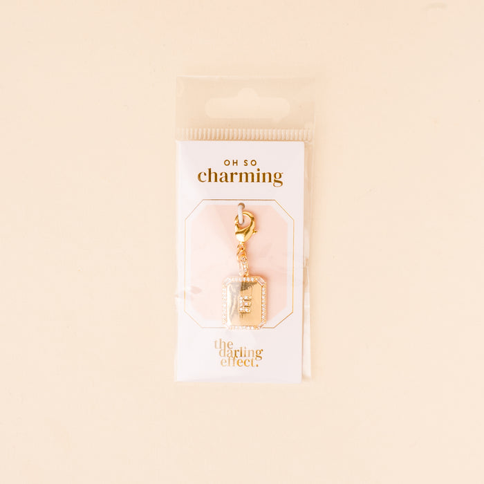 The E Jewelry Charm Initial Letter Gold by the-darling-effect features a gold-plated brass book-shaped charm with silver accents and a lobster clasp, presented on a white "OH SO charming" card in clear plastic packaging.