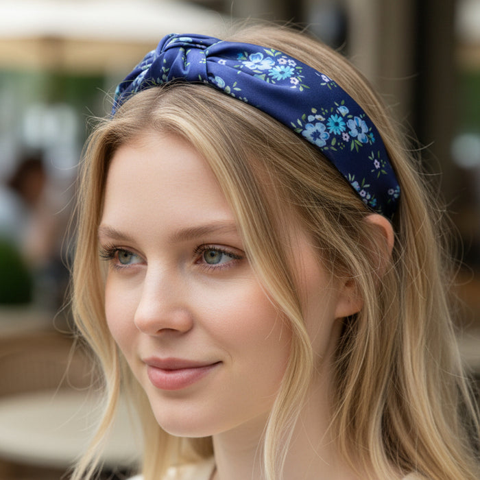 A young woman with blonde hair smiles softly outdoors, wearing The Darling Effect’s Endless Daydream Navy Classic Twist Headband. Blurred tables and chairs are visible in the background.