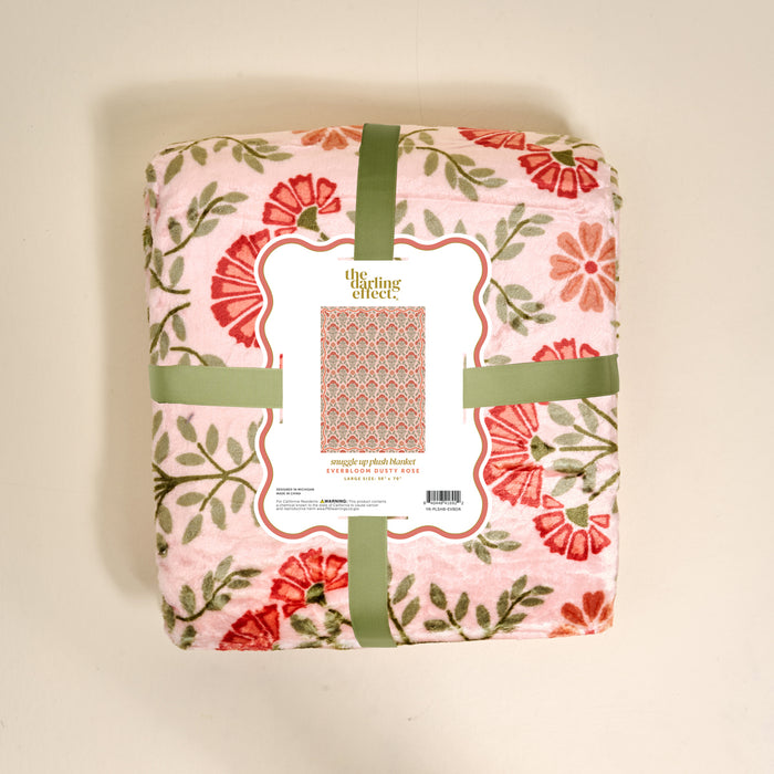 A folded pink floral Everbloom Dusty Rose Snuggle Up Plush Blanket by The Darling Effect, wrapped in green ribbon with a label showing red and green flowers and reading "snuggle up plush blanket.