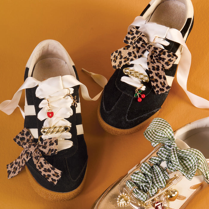 Three stylish sneakers with decorative bows, charms, and durable shoelaces: one black pair with cream and leopard-print bows, gold chain, and cherry charms; one gold pair with The Darling Effect Gingham Green Step It Up Shoelaces and gold charms on orange background.
