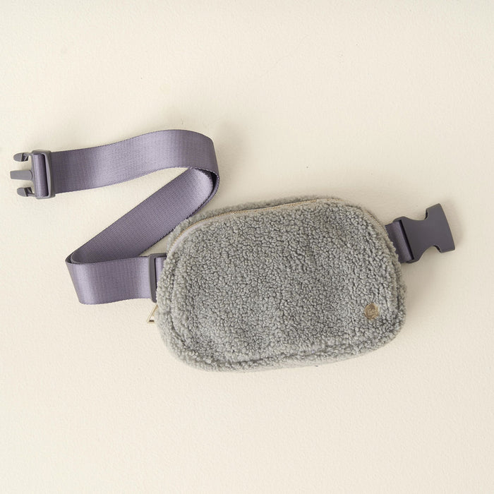 The Darling Effect Grey Sherpa Belt Bag is a small, rectangular, gray sherpa fabric bag with an adjustable matching strap and plastic buckle, shown on a light surface.