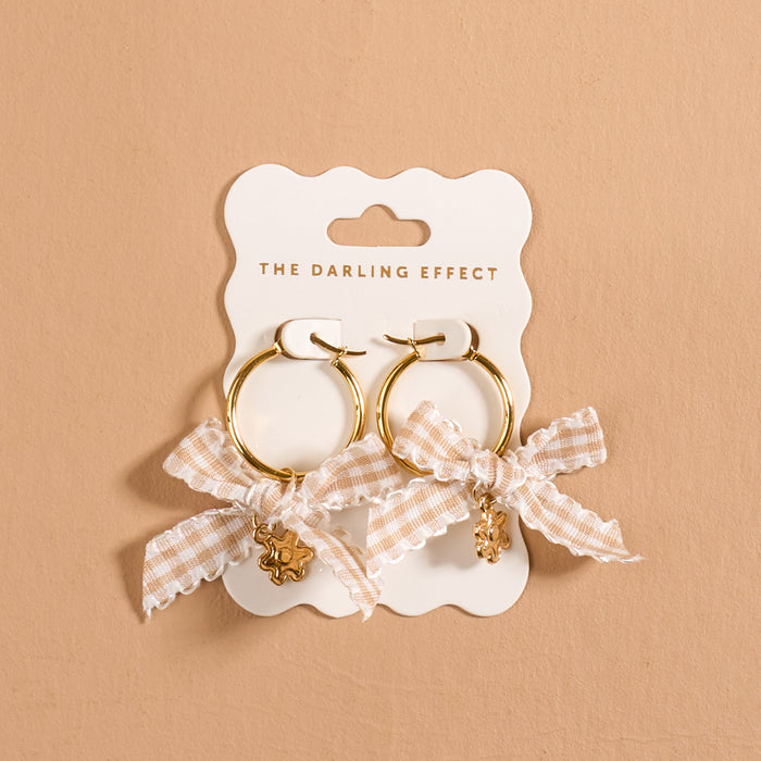 Harvest Honey Signature Hoop Earrings by The Darling Effect feature gold hoops adorned with checkered cream bows and delicate dangling charms, elegantly displayed on a branded card against a neutral beige background. These lightweight earrings beautifully combine playful elegance with modern charm, making them an ideal addition to any collection of charm earrings.