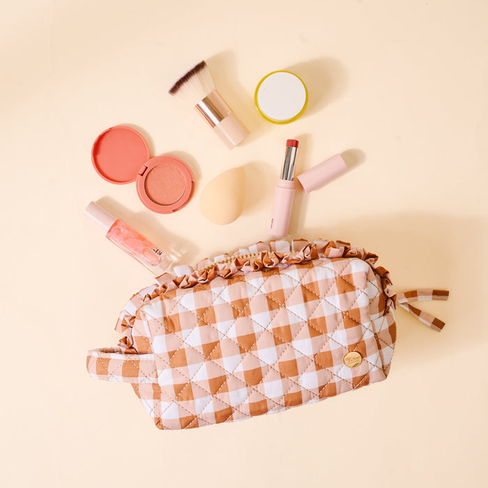 The Maple Gingham Essentials Quilted Cosmetic Pouch by The Darling Effect, adorned with a charming checkered pattern, is displayed on a light beige background surrounded by blush, lip gloss, a brush, blending sponge, and lipstick. Featuring a wipeable interior, this cosmetic bag effortlessly blends style with functionality.