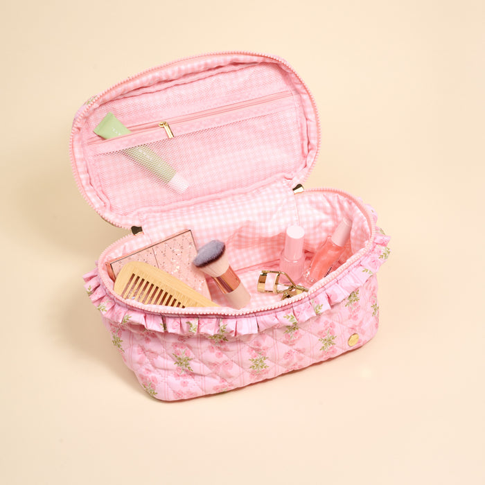 The Darling Effect’s Oh So Fancy Pink Glam & Go Quilted Cosmetic Bag, featuring floral patterns and ruffled edges, is open to reveal neatly arranged makeup brushes, lip products, a comb, and a tube on a light beige background.