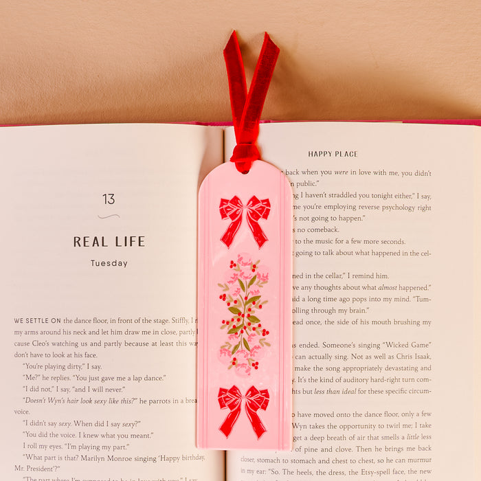 The Oh So Merry Acrylic Bookmark by The Darling Effect, featuring a vibrant floral design and a soft red velvet ribbon tassel, rests on an open book to mark the page titled REAL LIFE and HAPPY PLACE.