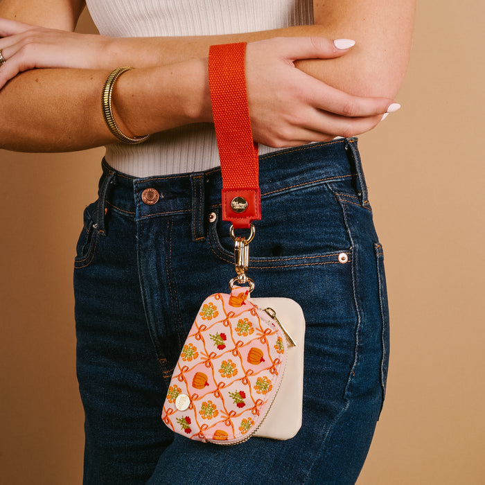 A person in a white sleeveless top and blue jeans holds The Darling Effect Patchwork Pumpkins Dynamic Duo Pouch Wristlet, featuring a red strap and clipped hands-free to a white item, perfect for organizing essentials against a beige background.