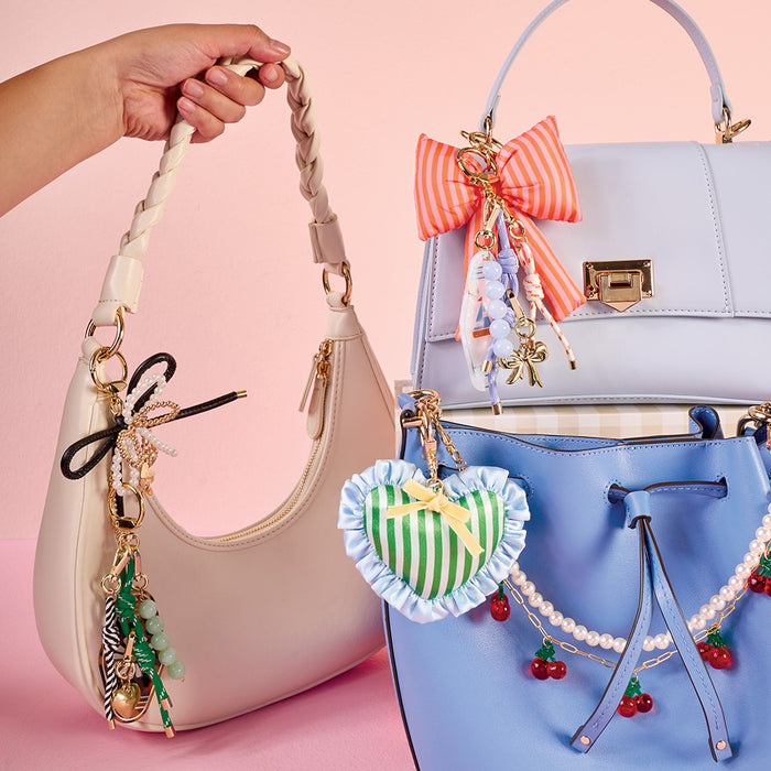 A hand holds a cream-colored shoulder bag with charms, beside a light purple handbag featuring The Darling Effect's Peachy Petals Puffy Bow Bag Charm and an orange bow charm, plus a blue bucket bag with heart and cherry charms.