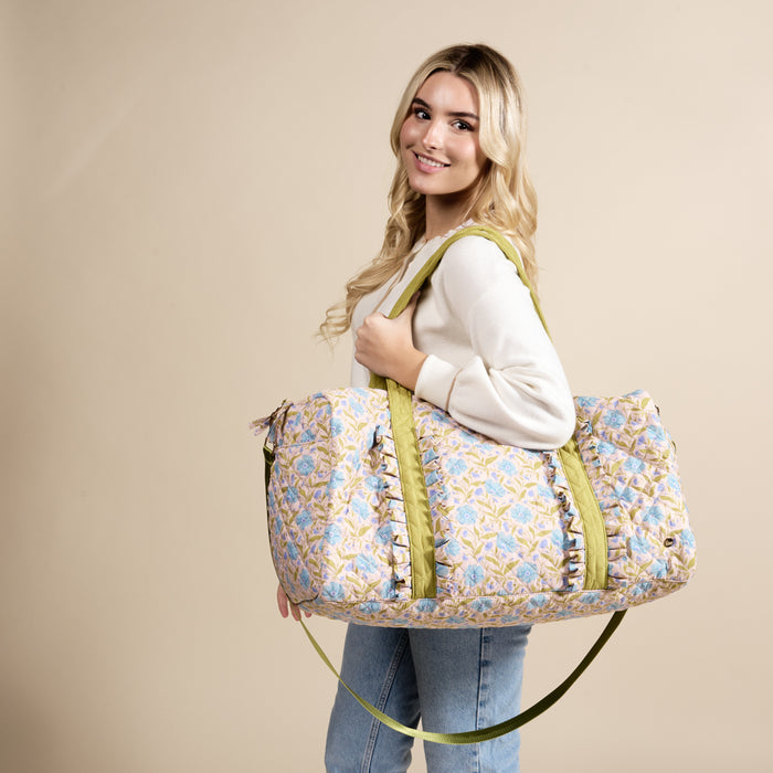 A smiling woman with long blonde hair holds The Darling Effect Poppin' Petals Aqua On-the-Go Quilted Weekender Bag, featuring green straps and an adjustable shoulder strap, against a plain beige background. She wears a light sweater and blue jeans.