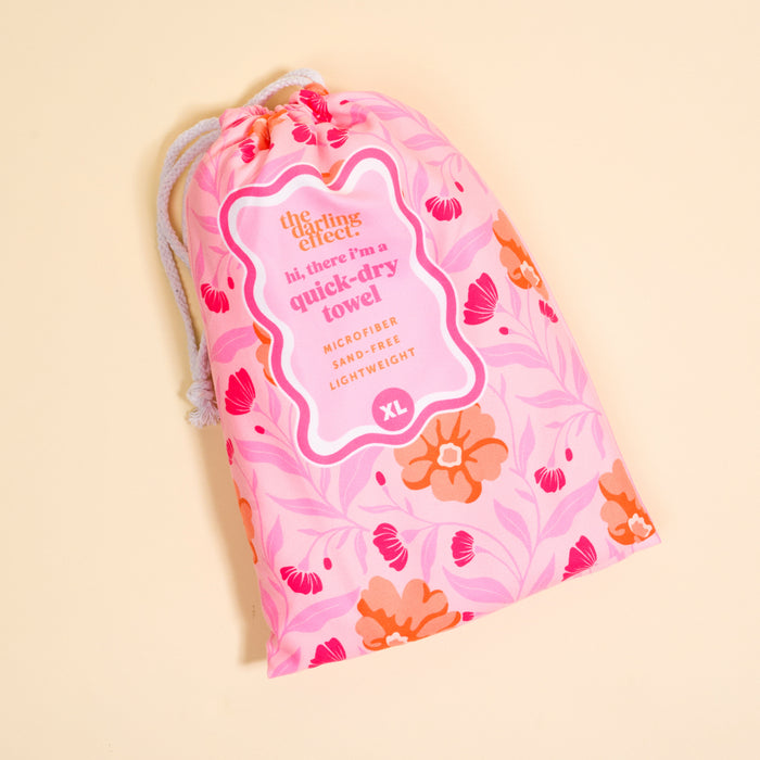 A pink drawstring bag with a floral pattern displays a label reading, “The Darling Effect Poppin' Petals Hot Pink XL Quick-Dry Beach Towel, microfiber, sand-free, lightweight,” on a pale yellow background.