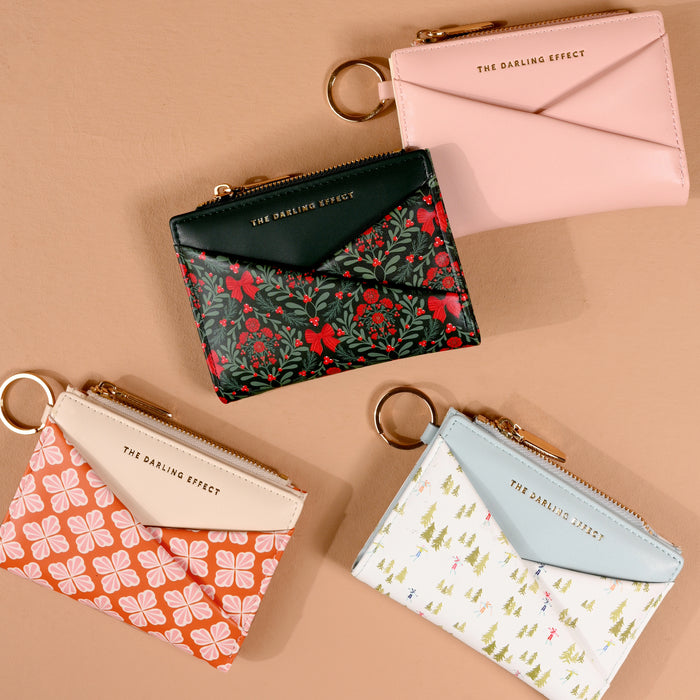 Four Sienna Spice Luxe Pocket Wallets by The Darling Effect are arranged on a tan surface. Each vegan leather wallet features an envelope flap, a keyring, and gold THE DARLING EFFECT text. Designs include floral, geometric, tree, criss-cross patterns, and a plain blush pink option.