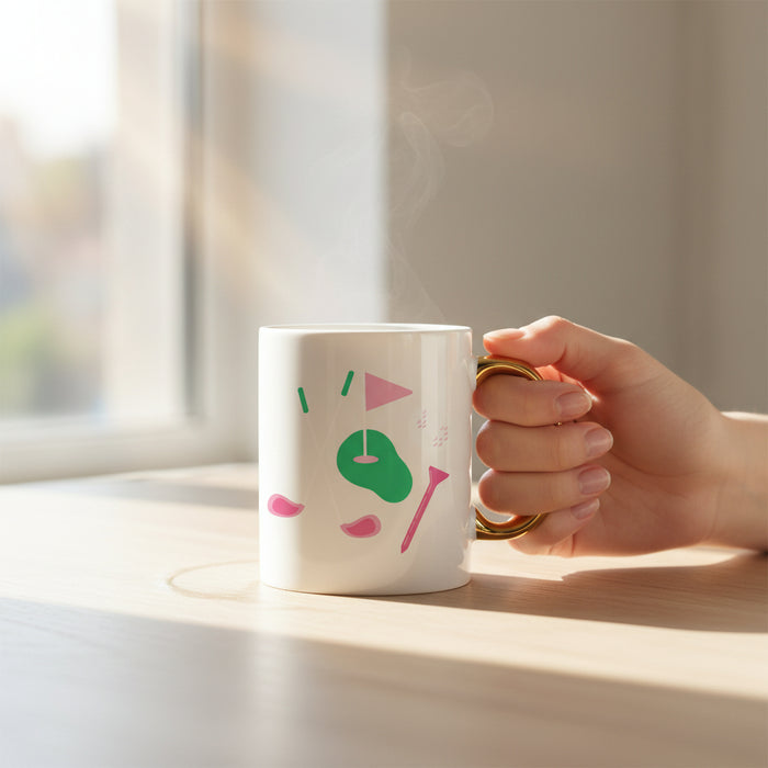 A hand holds The Golf Edit 11 oz Cozy Cute Ceramic Mug by The Darling Effect, with a gold handle and colorful design. Steam from coffee or tea rises as sunlight shines on the wooden surface.