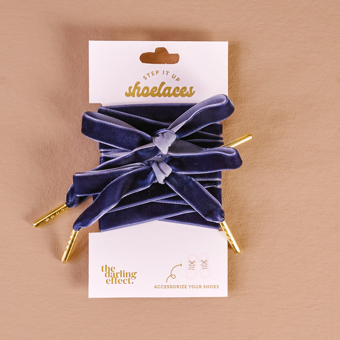 A pair of wide, stylish velvet blue shoelaces with gold tips are tied in bows and displayed on a branded card reading Velvet Blue Step It Up Shoelaces by The Darling Effect against a beige background.
