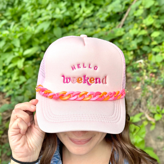 A person wears a light pink hat with "HELLO weekend" in colorful embroidery and The Darling Effect's Acrylic Sassy Trucker Hat Chain in pink and orange across the brim. Green leafy plants are blurred in the background.