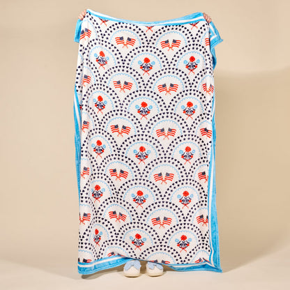 A person holds up The Darling Effect's All-American Darling Snuggle Up Plush Blanket, featuring red, white, and blue American flag and floral patterns with blue trim. Only their hands and shoes appear behind the soft blanket.