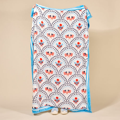 A person holds up The Darling Effect's All-American Darling Snuggle Up Plush Blanket, featuring red, white, and blue American flag and floral patterns with blue trim. Only their hands and shoes appear behind the soft blanket.