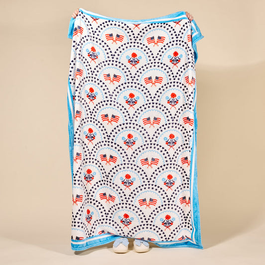 A person holds up The Darling Effect's All-American Darling Snuggle Up Plush Blanket, featuring red, white, and blue American flag and floral patterns with blue trim. Only their hands and shoes appear behind the soft blanket.