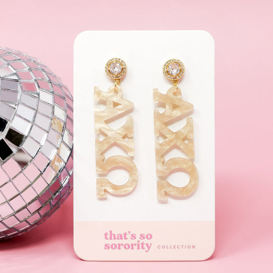 Alpha Chi Omega Sorority Letter Gold Acrylic Earrings by The Darling Effect, shown on a "that's so sorority collection" card beside a disco ball—lightweight and perfect for your bid-day basket, set against a pink backdrop.