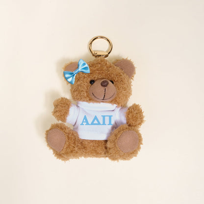 The Darling Effect's Alpha Delta Pi Teddy Bear Bag Charm is a small brown bear keychain with a blue bow and white shirt featuring blue ΑΔΠ Greek letters—perfect as a Bid Day gift or cute accessory.