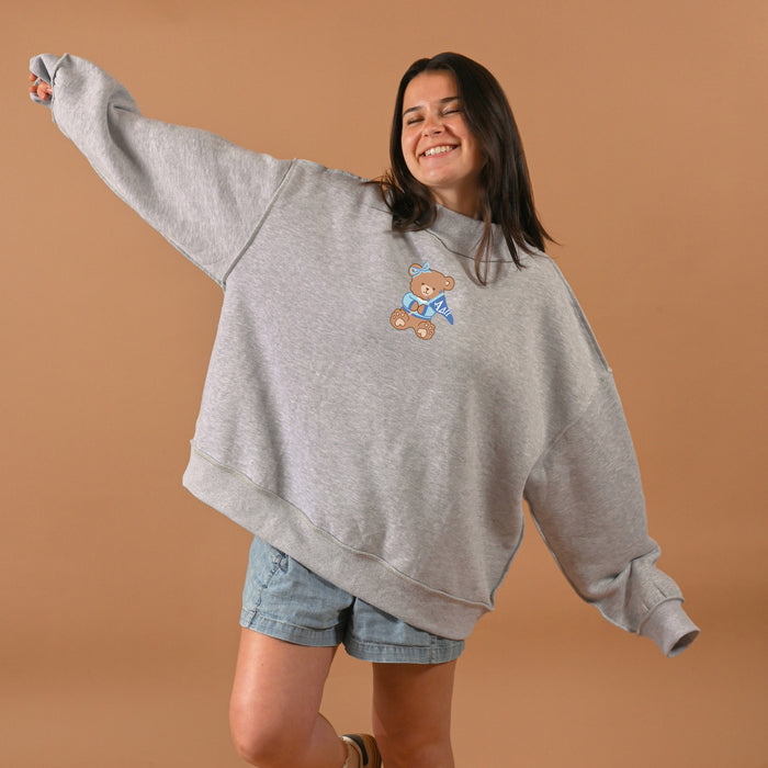 A woman smiles and extends her arms playfully while wearing The Darling Effect’s Alpha Delta Pi Teddy Bear Mockneck Sweatshirt in L/XL with denim shorts, standing against a tan background.