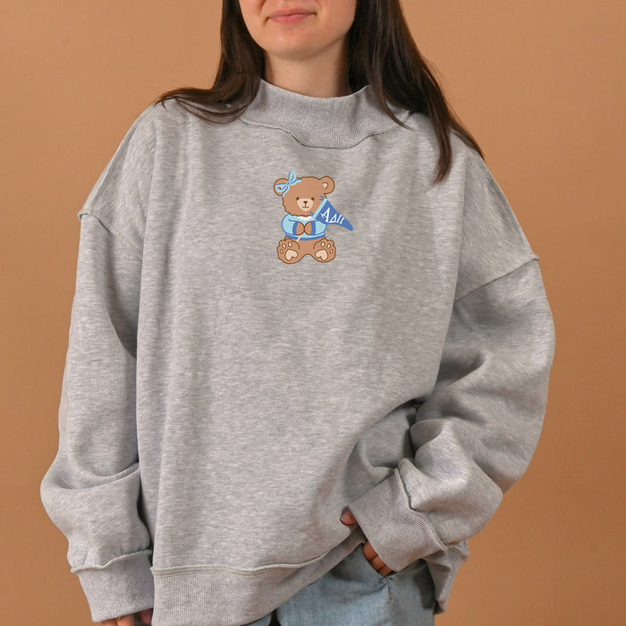 A woman wears The Darling Effect's Alpha Delta Pi Teddy Bear Mockneck Sweatshirt in size L/XL, featuring an oversized gray fit with a charming brown teddy bear graphic holding a blue flag that reads "Adil." Set against a solid beige background, her face is partially visible, and the cozy design evokes a classic sorority sweatshirt vibe.