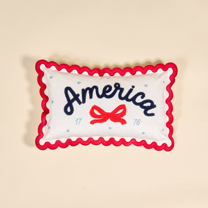 The Darling Effect's America Scalloped Rectangle Throw Pillow features white fabric, red wavy trim, "America" in blue script, a red bow, blue stars, and 1776. A charming piece for your collection, shown on a beige background.