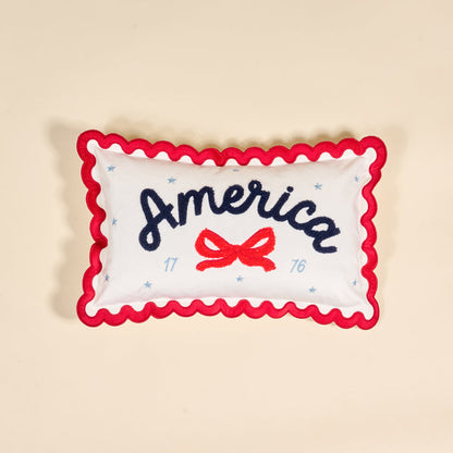 The Darling Effect's America Scalloped Rectangle Throw Pillow features white fabric, red wavy trim, "America" in blue script, a red bow, blue stars, and 1776. A charming piece for your collection, shown on a beige background.