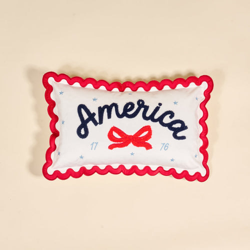 The Darling Effect's America Scalloped Rectangle Throw Pillow features white fabric, red wavy trim, "America" in blue script, a red bow, blue stars, and 1776. A charming piece for your collection, shown on a beige background.