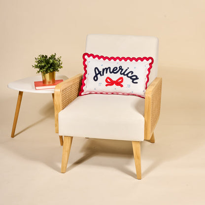 A modern cream armchair with wooden arms features The Darling Effect’s America Scalloped Rectangle Throw Pillow. Beside the chair, a white side table holds a potted plant and two stacked books.