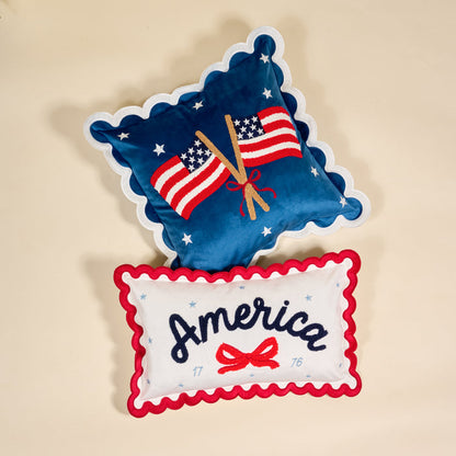 The Darling Effect's America Scalloped Rectangle Throw Pillow set features one blue pillow with embroidered flags and stars, and a white pillow with red trim, "America" text, a red bow, and 1776—perfect accent pieces.