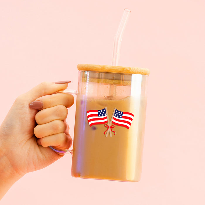 A hand holds The Darling Effect’s American Flags Square Glass Cup with bamboo lid and glass straw, featuring two small American flags crossed over a red ribbon, against a soft pink background.
