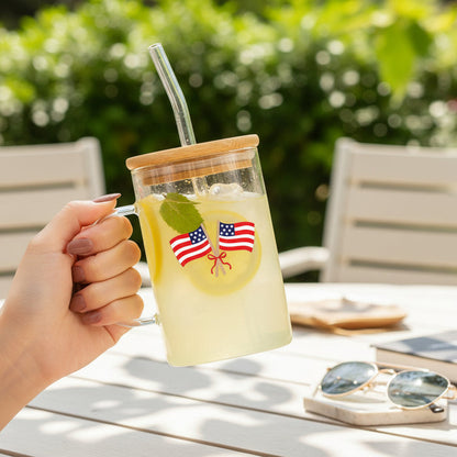 A hand holds The Darling Effect's American Flags Square Glass Cup with lemonade, ice, lemon slices, and mint. The cup features a bamboo lid, straw, and flag stickers. Sunglasses and a book sit on a white outdoor table in the background.