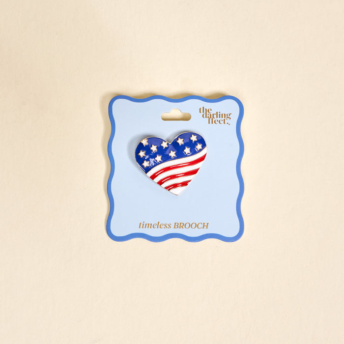 The Americana Heart Timeless Brooch by The Darling Effect features a heart-shaped American flag design with stars and stripes, presented on a light blue card—a vintage-inspired keepsake accessory.