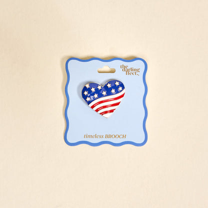 The Americana Heart Timeless Brooch by The Darling Effect features a heart-shaped American flag design with stars and stripes, presented on a light blue card—a vintage-inspired keepsake accessory.