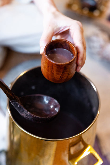 A hand holds a small wooden cup filled with a dark liquid above a large brass pot containing more of the same dark liquid, with a wooden spoon resting inside the pot.