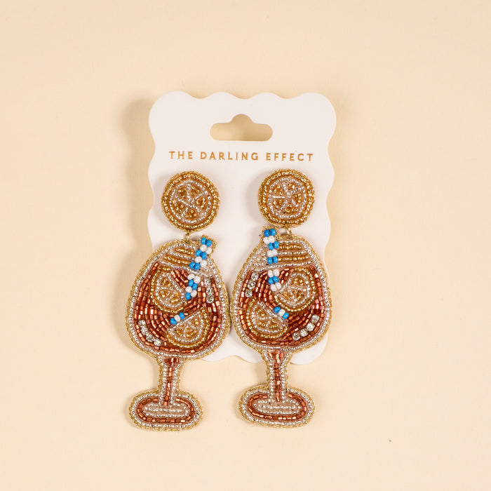 Aperol Spritz Beaded Earrings by The Darling Effect: vibrant, hand-beaded earrings shaped like wine glasses with gold, brown, blue, and white beads, displayed on a branded card against a beige background.