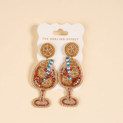 Aperol Spritz Beaded Earrings by The Darling Effect: vibrant, hand-beaded earrings shaped like wine glasses with gold, brown, blue, and white beads, displayed on a branded card against a beige background.