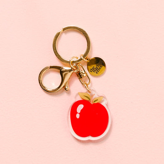 The Darling Effect’s Apple Acrylic Keychain features a durable red apple-shaped charm and a round “Critter Club” tag, displayed on a pale pink background.